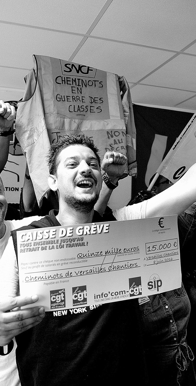 Militant souriant, poing levé, brandissant un chèque géant de caisse de grève de 15 000 € pour les cheminots de Versailles Chantiers, devant une banderole « Cheminots en guerre des classes ».